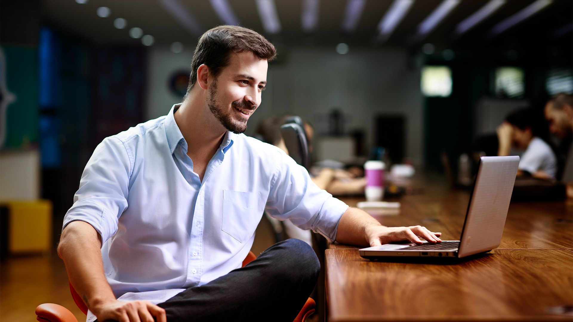 Um homem está sentado em seu escritório, usando um notebook. Ele está concentrado em seu trabalho, digitando com agilidade. O ambiente é iluminado e organizado. A cena transmite profissionalismo e foco.